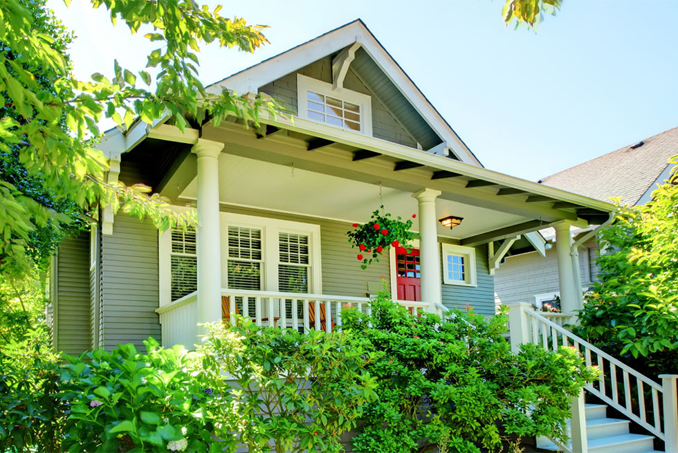 Front view of a charming gray house with white trim, a red door, a covered porch with white railings, surrounded by green bushes and trees on a sunny day
