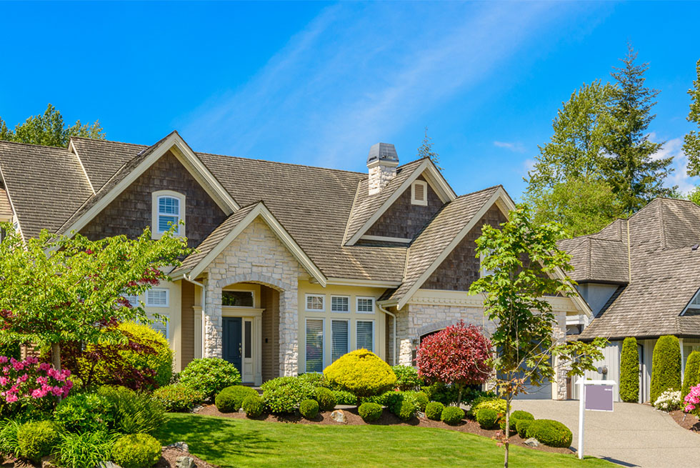 Large suburban house with stone and wood exterior, surrounded by well-maintained green shrubs, trees, and colorful flowering plants under a clear blue sky.