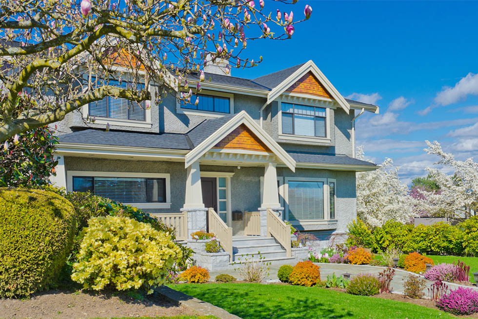 Two-story house with gray exterior and wooden accents, surrounded by a colorful garden with green shrubs, blooming flowers, and trees under a bright blue sky.