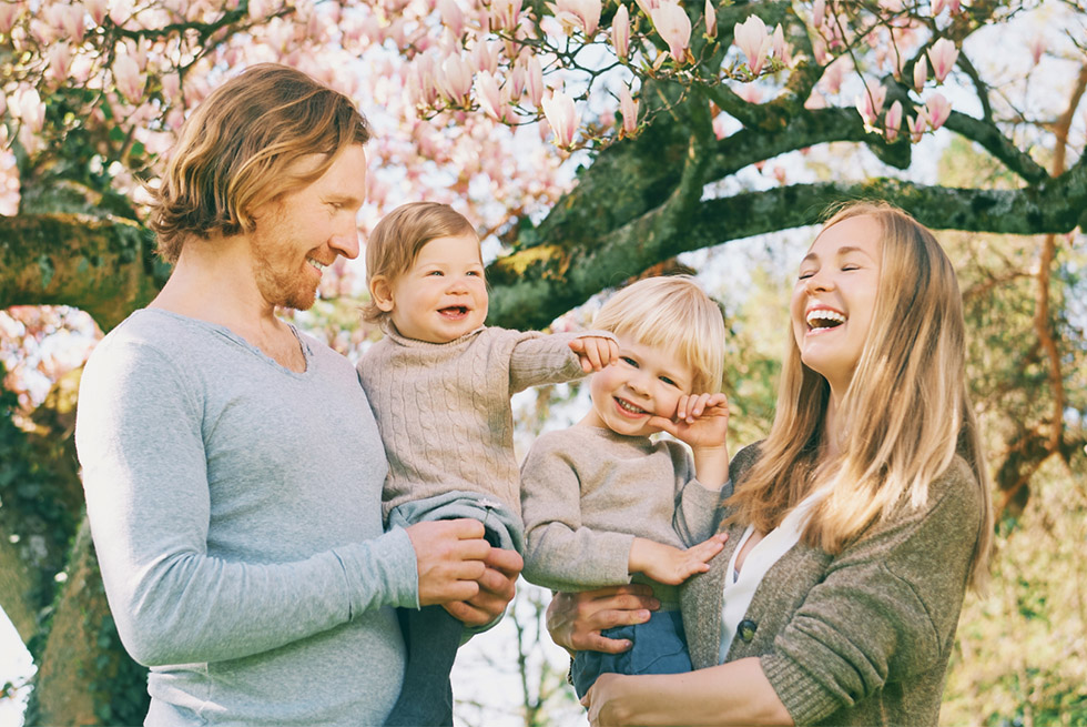 A family of four standing outdoors beneath flowering tree branches: a smiling man holds a laughing toddler while a small child