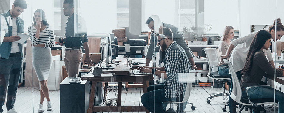 A modern office space with several people working on computers, collaborating, and standing while discussing work