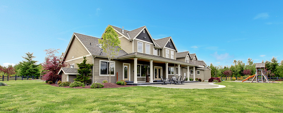 Spacious modern two-story house with large porch and outdoor seating, surrounded by green lawn and trees under a clear blue sky, with a children's playset in the background.