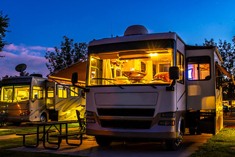 Exterior view of two illuminated RVs parked at a campsite during twilight, with picnic tables nearby and trees in the background. Exterior view of two illuminated RVs parked at a campsite during twilight, with picnic tables nearby and trees in the background.