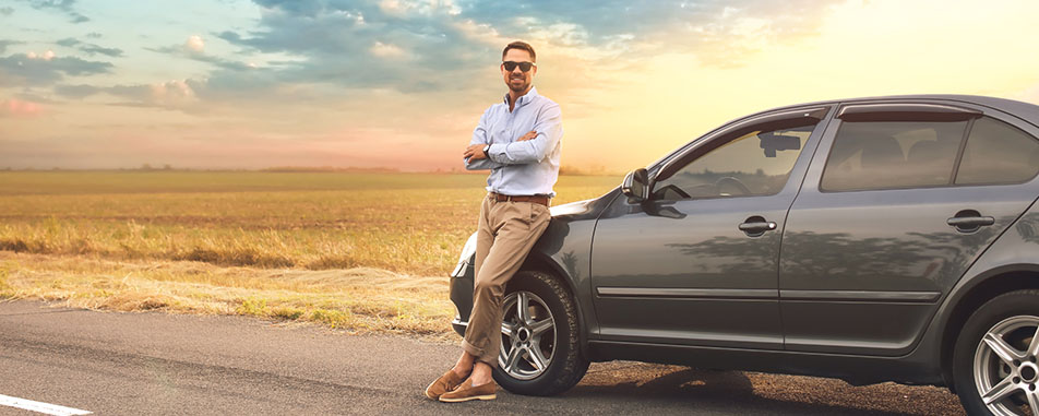 Man standing with arms crossed, leaning against a dark-colored car parked on the side of a road, with a sunset and open field in the background.