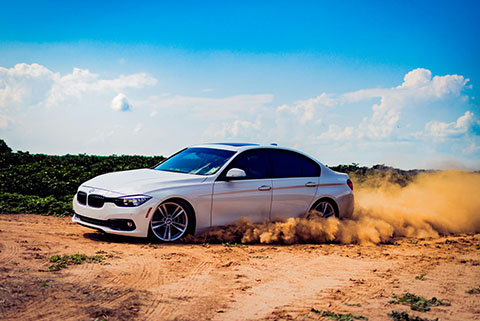 White BMW sedan drifting on a dirt road kicking up a cloud of dust under a bright blue sky with scattered clouds