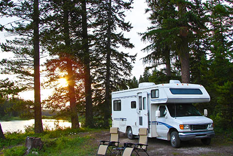 White camper van parked in a forested area near a lake at sunset with two folding chairs set up outside